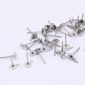 May include: A collection of silver-colored earring posts with flat, round pads and small loops. The posts are scattered across a white surface, showcasing their simple, minimalist design. These components are ideal for DIY jewelry making.