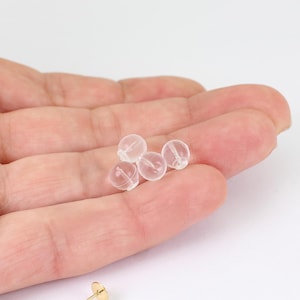 May include: A close-up of five small, clear, round earring backs. The earring backs are made of a translucent material and are resting in the palm of a hand. The background is white.