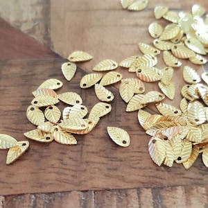 May include: A collection of small, gold-colored leaf charms scattered on a wooden surface. Each charm has a detailed leaf vein pattern and a small hole for stringing. The charms are likely for jewelry making or other crafts.