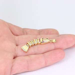 May include: A gold-colored pendant with a segmented design, adorned with red, clear, and green gemstones. The pendant has a small loop for a chain and is held in a person's hand against a white background. The pendant's design is reminiscent of a stylized fish.