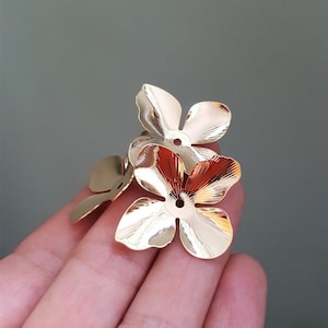 May include: Three gold-coloured metal flower shapes with a hole in the centre. The flowers are arranged in a cluster and are being held in a hand.