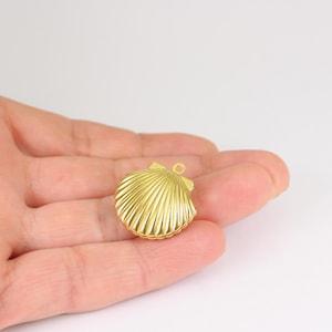 May include: A gold-colored seashell charm with a loop for attaching to jewelry. The charm has a detailed, ridged surface and a small loop at the top. The seashell is held in a person's hand against a white background.