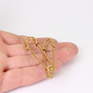 May include: Two gold-colored safety pins. The pins are made of a shiny metal and have a classic design with a looped head and a pointed end. The pins are shown against a white background.