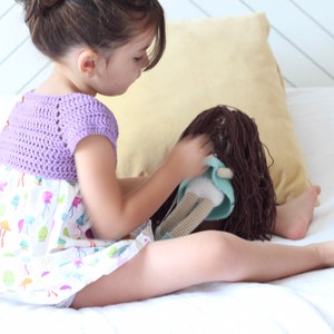 Puede incluir: Una niña pequeña con un top de crochet morado y un vestido blanco con estampado de medusas juega con una muñeca de pelo castaño en una cama blanca.