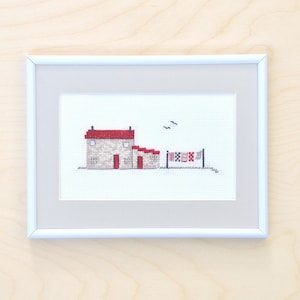 Puede incluir: Una bordadura de punto de cruz enmarcada que representa una pequeña casa con un techo rojo y una puerta roja. La casa está rodeada de una pequeña valla con banderas rojas y blancas colgando de ella. Hay dos pájaros volando en el cielo sobre la casa.