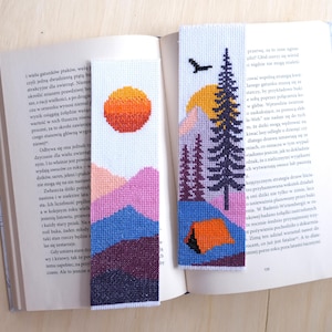May include: Two rectangular cross-stitch bookmarks featuring outdoor scenes. One displays a sunset over layered purple and blue mountains. The other shows a tall pine tree, a bird in flight, a yellow sun, and an orange tent on a blue landscape.