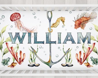 May include: A white crib sheet featuring an underwater theme with a jellyfish, seahorse, and lionfish. The name "WILLIAM" is displayed in teal letters, surrounded by an anchor, coral, and seaweed.
