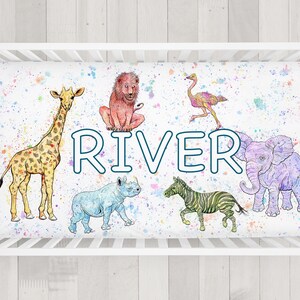 May include: A fitted crib sheet featuring watercolor illustrations of a giraffe, lion, ostrich, rhino, zebra, and elephant. The name "RIVER" is displayed in blue letters. The sheet has a white background with colorful paint splatters.