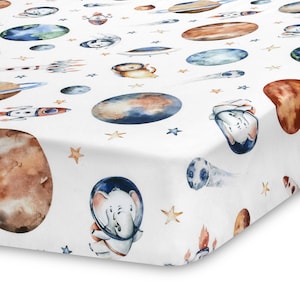 May include: White fitted crib sheet with a space-themed print. The print features planets, stars, rockets, and animals wearing astronaut suits.