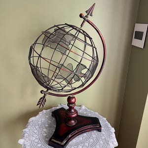 May include: A wire globe with a wooden base and an arrow pointing up. The globe is on a white doily on a wooden table.