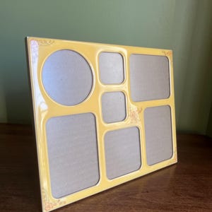 May include: A yellow multi-photo frame with a glossy finish. The frame has a variety of openings, including a large circle, squares, and rectangles. Floral accents are visible in the corners. The frame is displayed on a wooden surface.