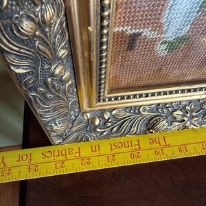 May include: A framed needlepoint artwork featuring a textured brown background and light-coloured stitched details, encased in an ornate, dark gold-toned frame with floral relief patterns, positioned above a yellow ruler showing 17 to 25 inches.