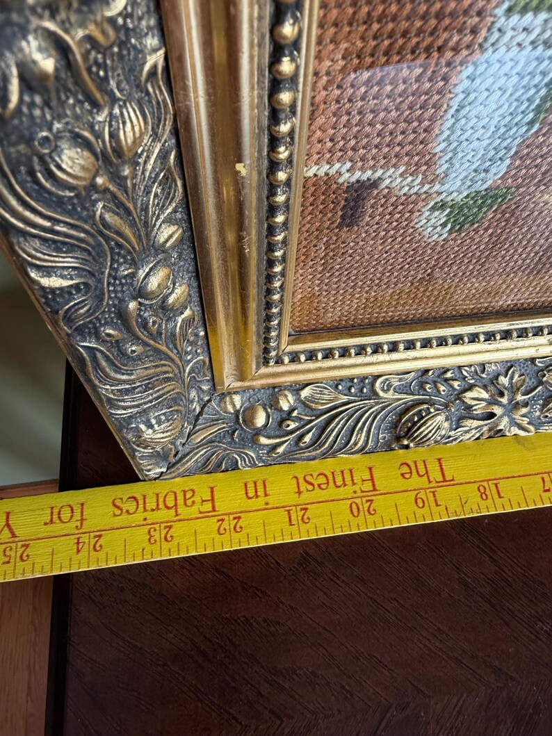 May include: A framed needlepoint artwork featuring a textured brown background and light-coloured stitched details, encased in an ornate, dark gold-toned frame with floral relief patterns, positioned above a yellow ruler showing 17 to 25 inches.