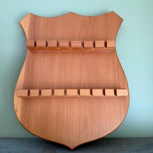 May include: Wooden spoon rack in a shield shape. The rack is made of light brown wood and features two rows of rectangular slots for holding spoons. The shield has a decorative top and a pointed bottom.