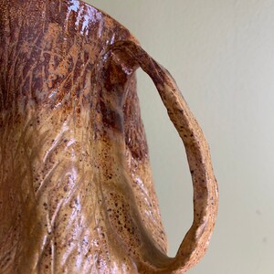 May include: A brown ceramic mug with a textured, tree bark-like design. The mug has a large, rounded handle.