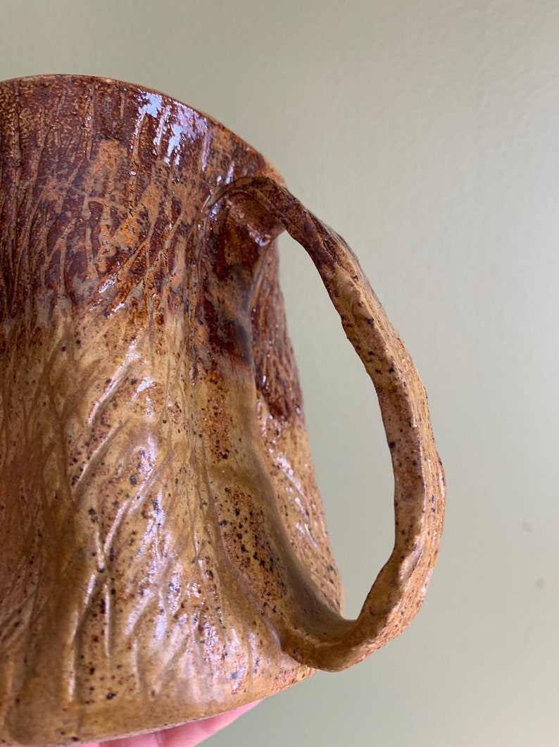 May include: A brown ceramic mug with a textured, tree bark-like design. The mug has a large, rounded handle.