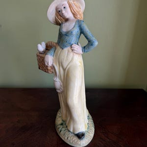 May include: A porcelain figurine of a person wearing a wide-brimmed hat, a blue long-sleeved top, and a long yellow skirt. The figure holds a brown woven basket containing two small white birds and stands on a textured oval base.