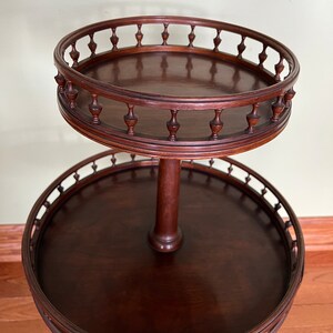May include: A two-tiered wooden serving table with a dark brown finish. The top tier has a decorative, scalloped edge with a series of small, turned wooden finials. The bottom tier has a similar scalloped edge with turned wooden finials.