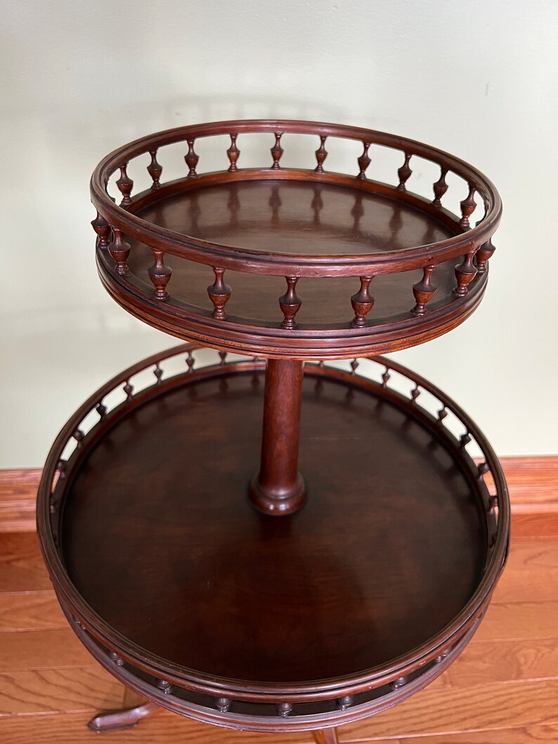 May include: A two-tiered wooden serving table with a dark brown finish. The top tier has a decorative, scalloped edge with a series of small, turned wooden finials. The bottom tier has a similar scalloped edge with turned wooden finials.