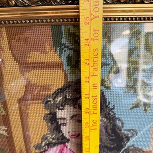 May include: A framed needlepoint tapestry depicting a person with dark hair wearing a pink and white garment, positioned next to a yellow measuring tape showing a length of approximately 30 cm from the 38 to 69 cm marks on the tape.