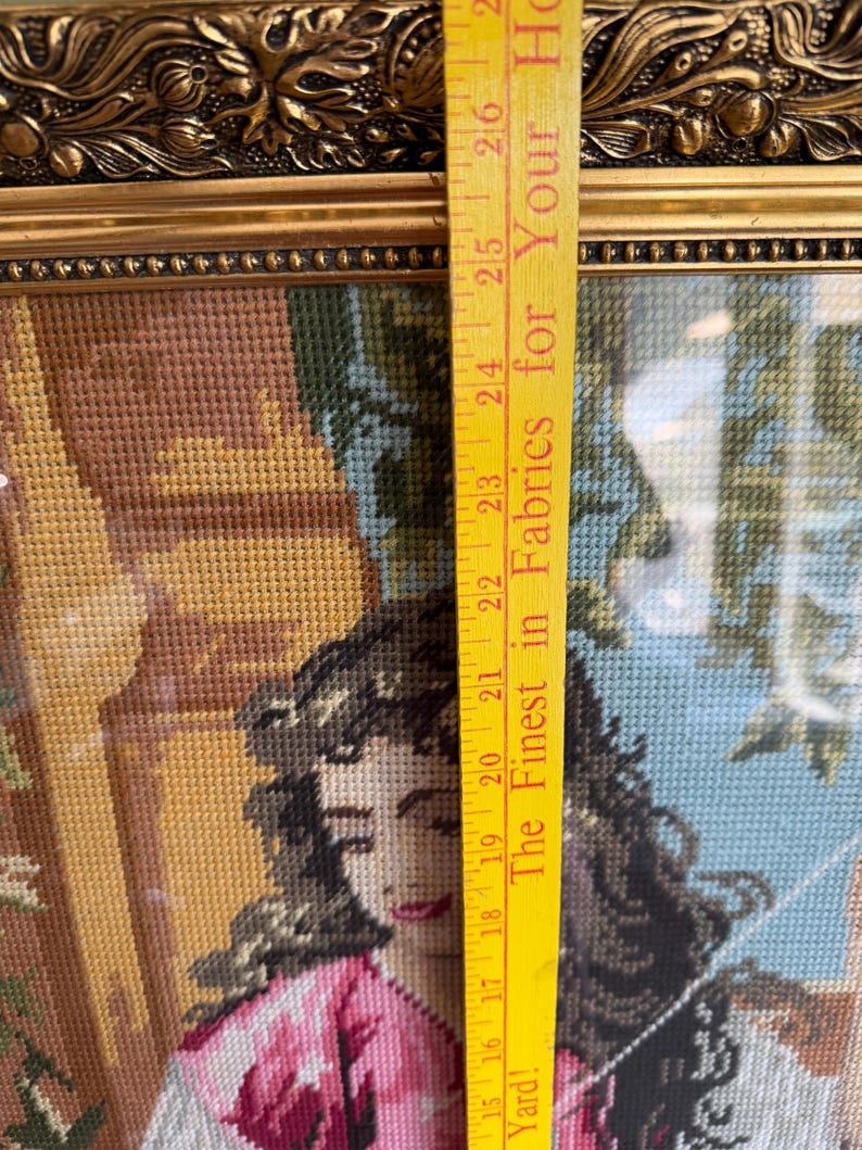 May include: A framed needlepoint tapestry depicting a person with dark hair wearing a pink and white garment, positioned next to a yellow measuring tape showing a length of approximately 30 cm from the 38 to 69 cm marks on the tape.