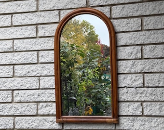 May include: An arched wall mirror featuring a medium-brown wooden frame with a rounded top and straight sides. The mirror glass reflects a view of green trees and foliage, and the base of the frame is accented with thin, wrapped rattan details.