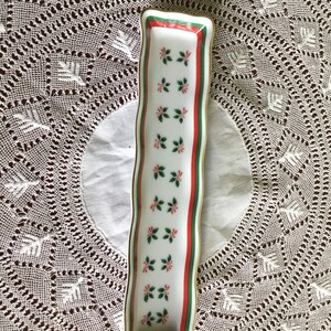 May include: A white rectangular ceramic dish with a gold rim. The dish has a green and red border with a repeating pattern of green leaves and red flowers.