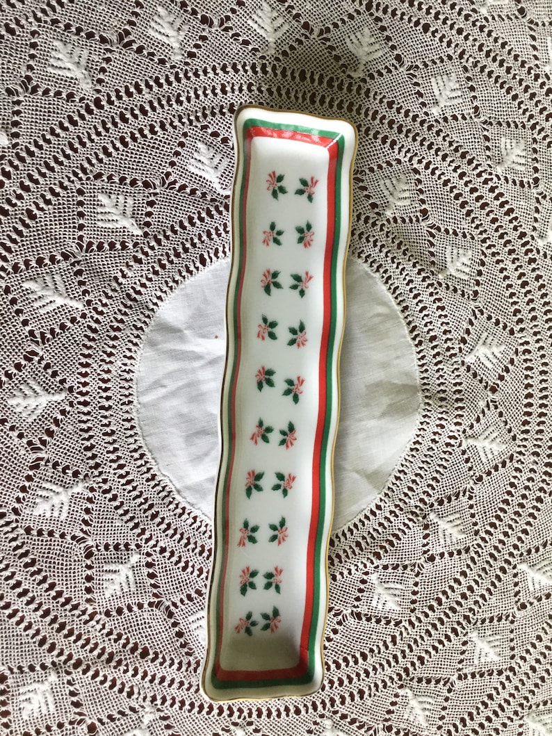 May include: A white rectangular ceramic dish with a gold rim. The dish has a green and red border with a repeating pattern of green leaves and red flowers.