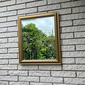 Puede incluir: Un espejo rectangular con marco dorado que refleja un exuberante jardín verde y un cielo nublado. El espejo está montado en una pared de ladrillos gris claro, creando un contraste entre el marco ornamentado y el fondo texturizado.