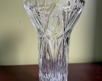 May include: A clear, cut-glass vase with a flared top and a cylindrical body. The vase features intricate geometric patterns and vertical lines, creating a textured appearance. The vase is designed to hold flowers and is suitable for home decor.