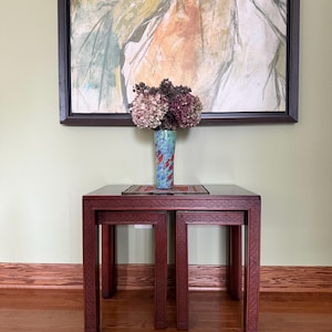 May include: A set of dark wood nesting tables with intricate carved geometric patterns on the legs and apron. A tall, cylindrical, multi-colored ceramic vase with blue, red, and green patterns holds a bouquet of dried hydrangea flowers.