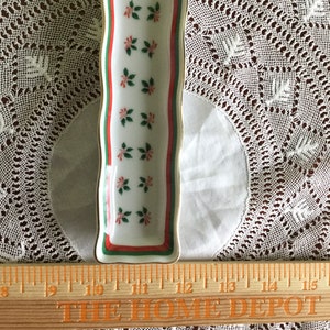 May include: A white ceramic serving dish with a red, green, and gold trim. The dish is decorated with a repeating pattern of green leaves and red berries. The dish is 12 inches long.