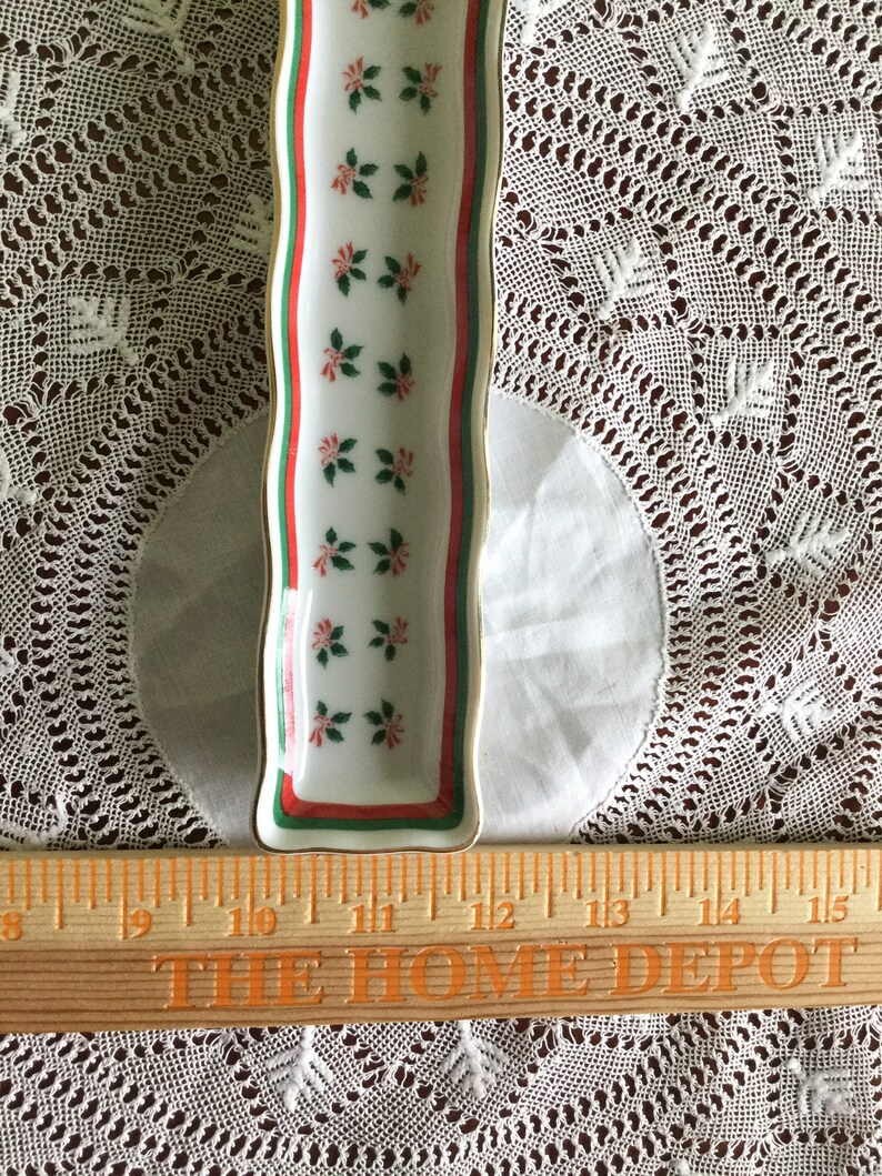 May include: A white ceramic serving dish with a red, green, and gold trim. The dish is decorated with a repeating pattern of green leaves and red berries. The dish is 12 inches long.