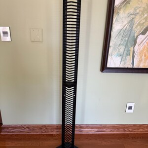 Peut inclure: Une sculpture décorative en métal noir de grande taille, avec un design géométrique. La sculpture a une base carrée et une série de découpes horizontales et verticales. Le sommet de la sculpture a une forme triangulaire. La sculpture est posée sur un sol en bois.