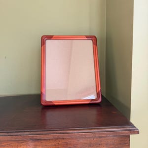 May include: A rectangular picture frame featuring a smooth, glossy finish with a gradient color scheme transitioning from bright orange at the top to a deep, dark reddish-brown at the bottom corners, enclosing a plain, light beige backing board.