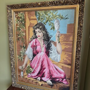 May include: A framed needlepoint tapestry depicting a person with long dark hair wearing a pink dress and white sleeves, sitting on a stone ledge while holding a spindle and thread. The artwork is encased in an ornate, textured gold-toned frame.