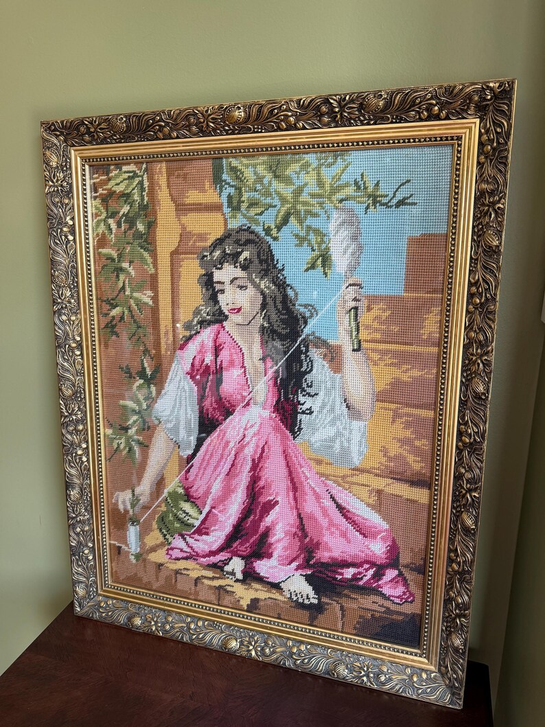 May include: A framed needlepoint tapestry depicting a person with long dark hair wearing a pink dress and white sleeves, sitting on a stone ledge while holding a spindle and thread. The artwork is encased in an ornate, textured gold-toned frame.