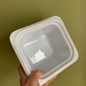 Puede incluir: Un plato cuadrado de cerámica blanca con un borde elevado. El plato está vacío y tiene un acabado brillante.