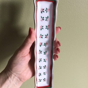 May include: A white ceramic trinket tray with a red and green floral design and a gold trim. The tray is rectangular and has a wavy edge.