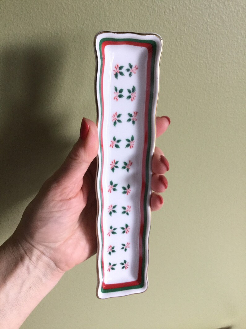 May include: A white ceramic trinket tray with a red and green floral design and a gold trim. The tray is rectangular and has a wavy edge.