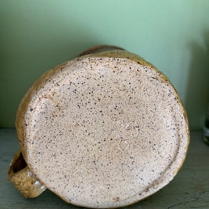 May include: The bottom of a ceramic mug with a speckled brown and tan glaze. The mug has a handle and a rounded bottom.