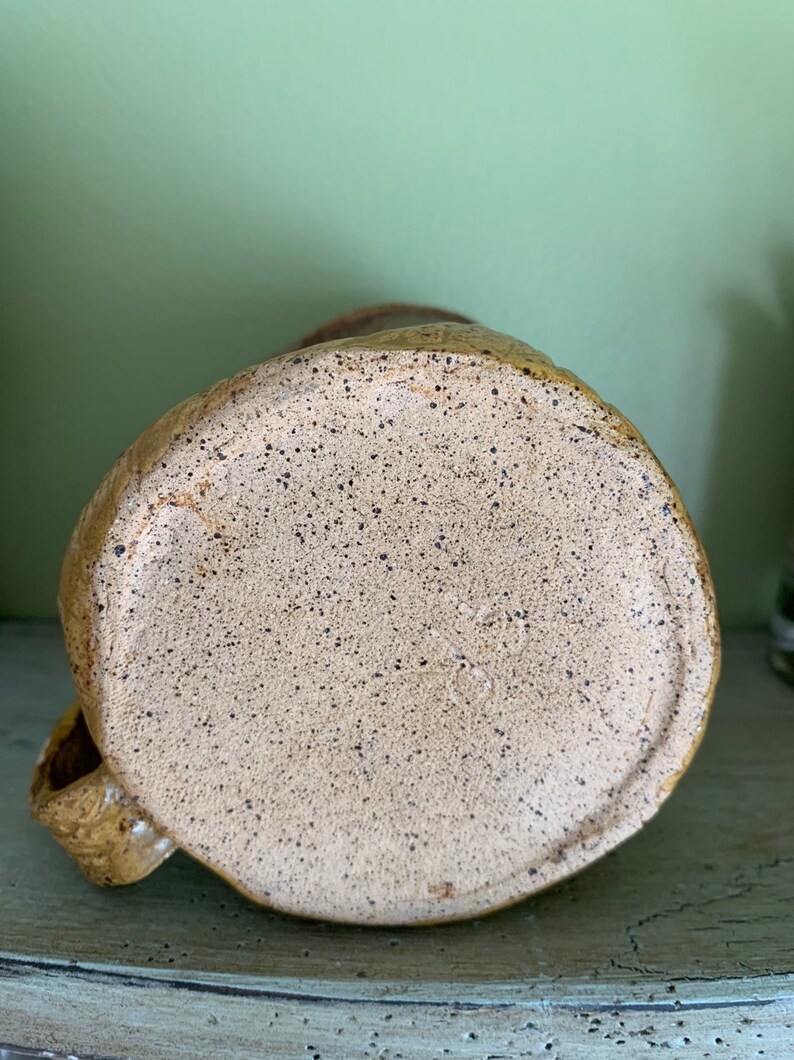 May include: The bottom of a ceramic mug with a speckled brown and tan glaze. The mug has a handle and a rounded bottom.