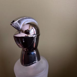 Pode incluir: Uma garrafa decorativa com uma rolha prateada em forma de capacete de cavaleiro. A garrafa tem um aspeto de vidro fosco, e o capacete é altamente reflexivo, com detalhes intrincados. O design evoca um tema medieval.