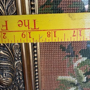 May include: A framed needlepoint tapestry featuring a floral pattern in shades of green and brown, set within an ornate gold-coloured frame. A yellow ruler is positioned across the frame, showing a measurement between 43 and 51 centimetres.