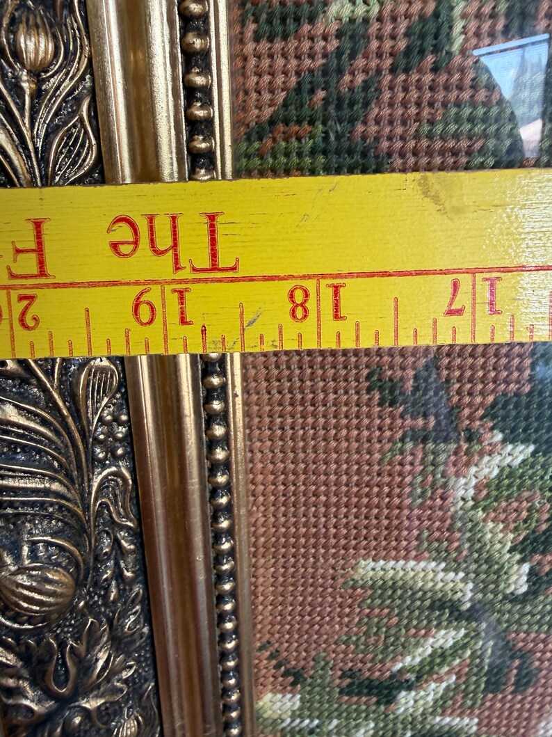 May include: A framed needlepoint tapestry featuring a floral pattern in shades of green and brown, set within an ornate gold-coloured frame. A yellow ruler is positioned across the frame, showing a measurement between 43 and 51 centimetres.