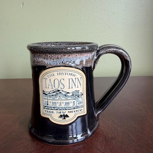 May include: A black ceramic mug with a handle, featuring a label that reads "The Historic Taos Inn, Taos, New Mexico." The mug has a speckled brown rim and a glossy finish. The label depicts a building and mountain scene.