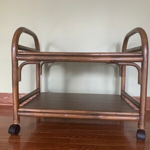 May include: A two-tiered wooden serving cart with a brown finish. The cart has a rectangular top and bottom shelf and is supported by four wheels. The cart is made of natural wood with a dark brown stain.