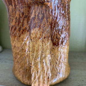 May include: A ceramic mug with a textured, tree bark-like design in shades of brown and yellow. The mug has a large handle.