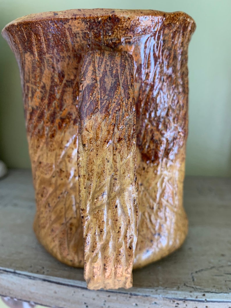 May include: A ceramic mug with a textured, tree bark-like design in shades of brown and yellow. The mug has a large handle.