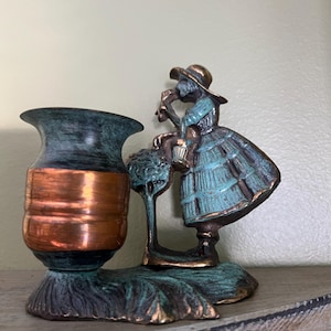 Peut inclure: Une figurine en bronze vintage d'une femme en robe bleue tenant un arrosoir, avec un vase de couleur cuivre fixé à la base. La figurine a une finition patine verte.